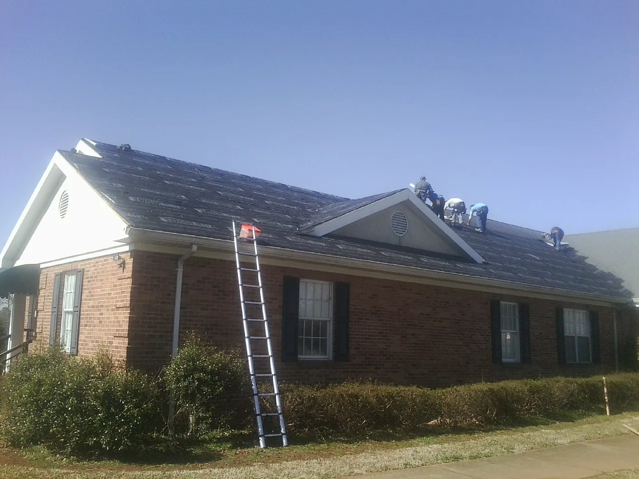 Crew tearing shingles off a brick ranch roof with a ladder set — Metal Roof Repair work by our crew in Rose Creek