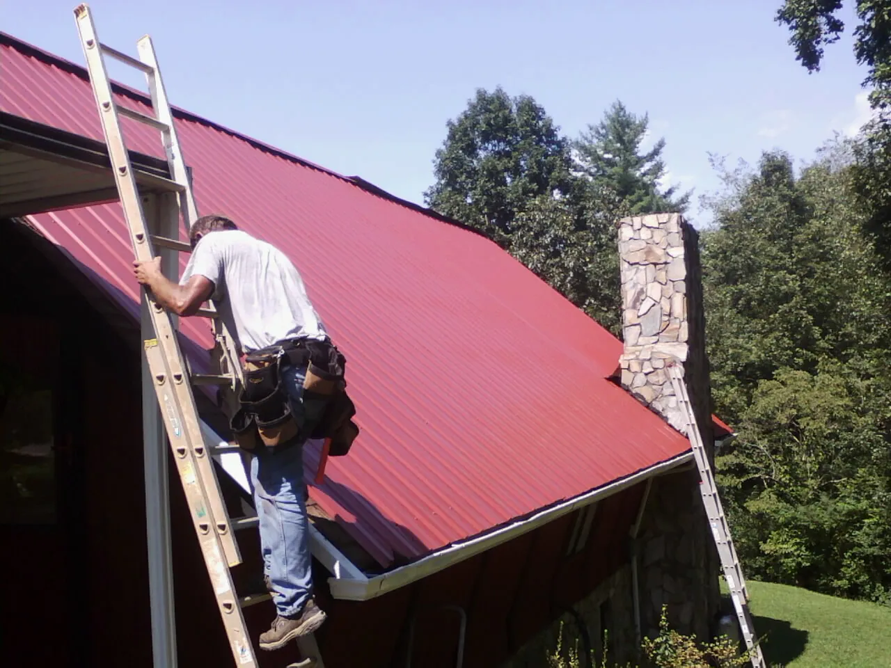 Roofer on a ladder beside a red metal roof and stone chimney — Soffit & Fascia Repair by Roofing Brothers in Rose Creek